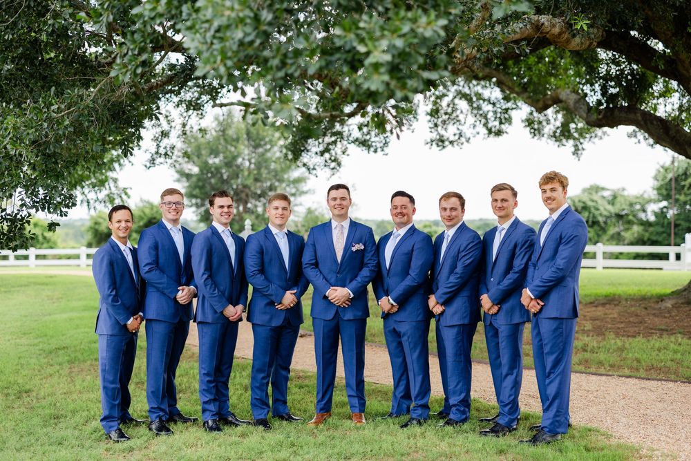 A wedding party of groomsmen in matching blue suits pose outdoors under green trees.