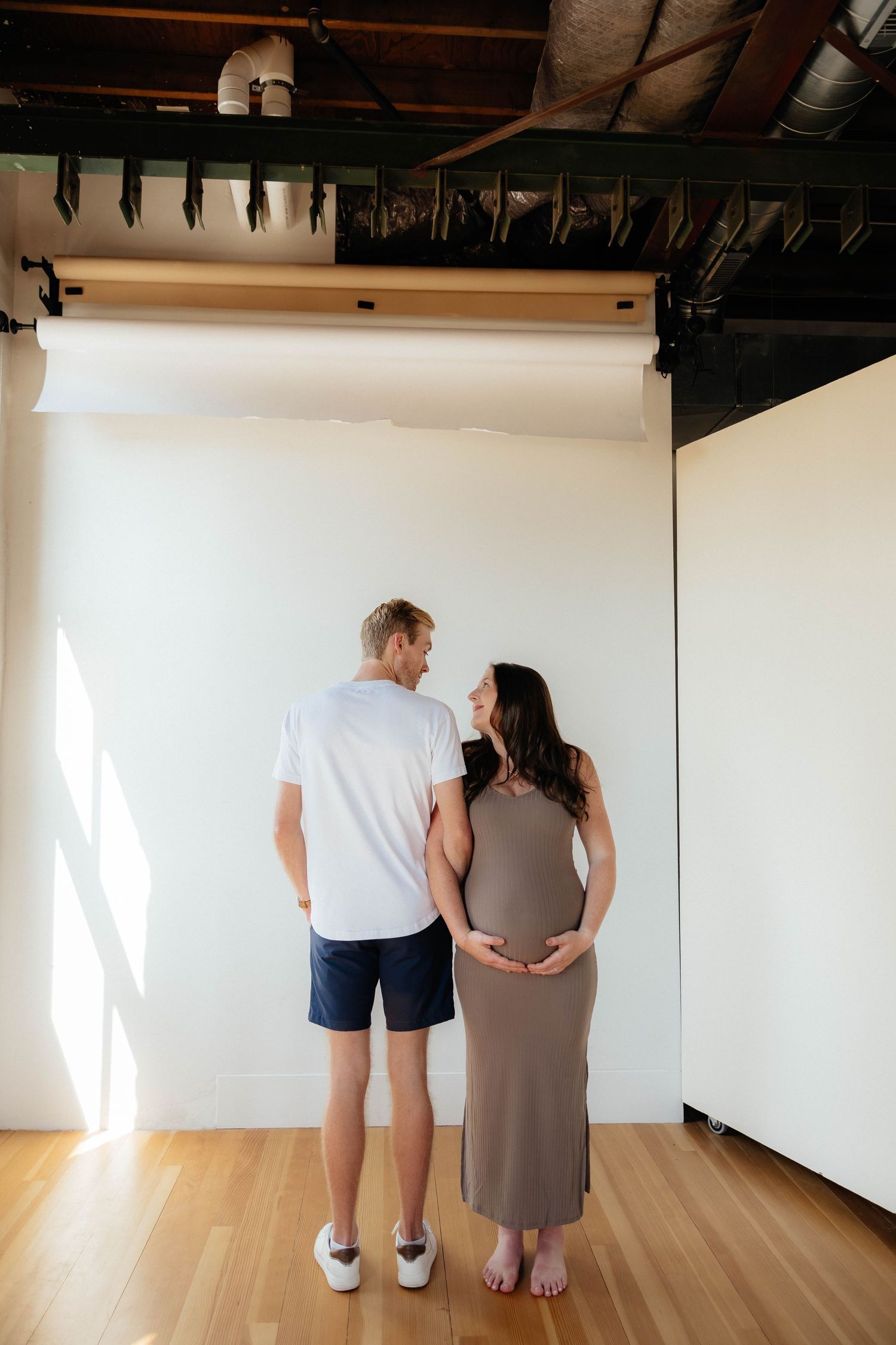 A woman in a brown dress and a man in a white shirt stand together on hardwood flooring against a white wall.