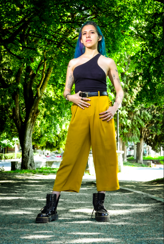 A person wearing combat boots, yellow wide-leg pants and black one-shoulder top stands confidently in a park setting.