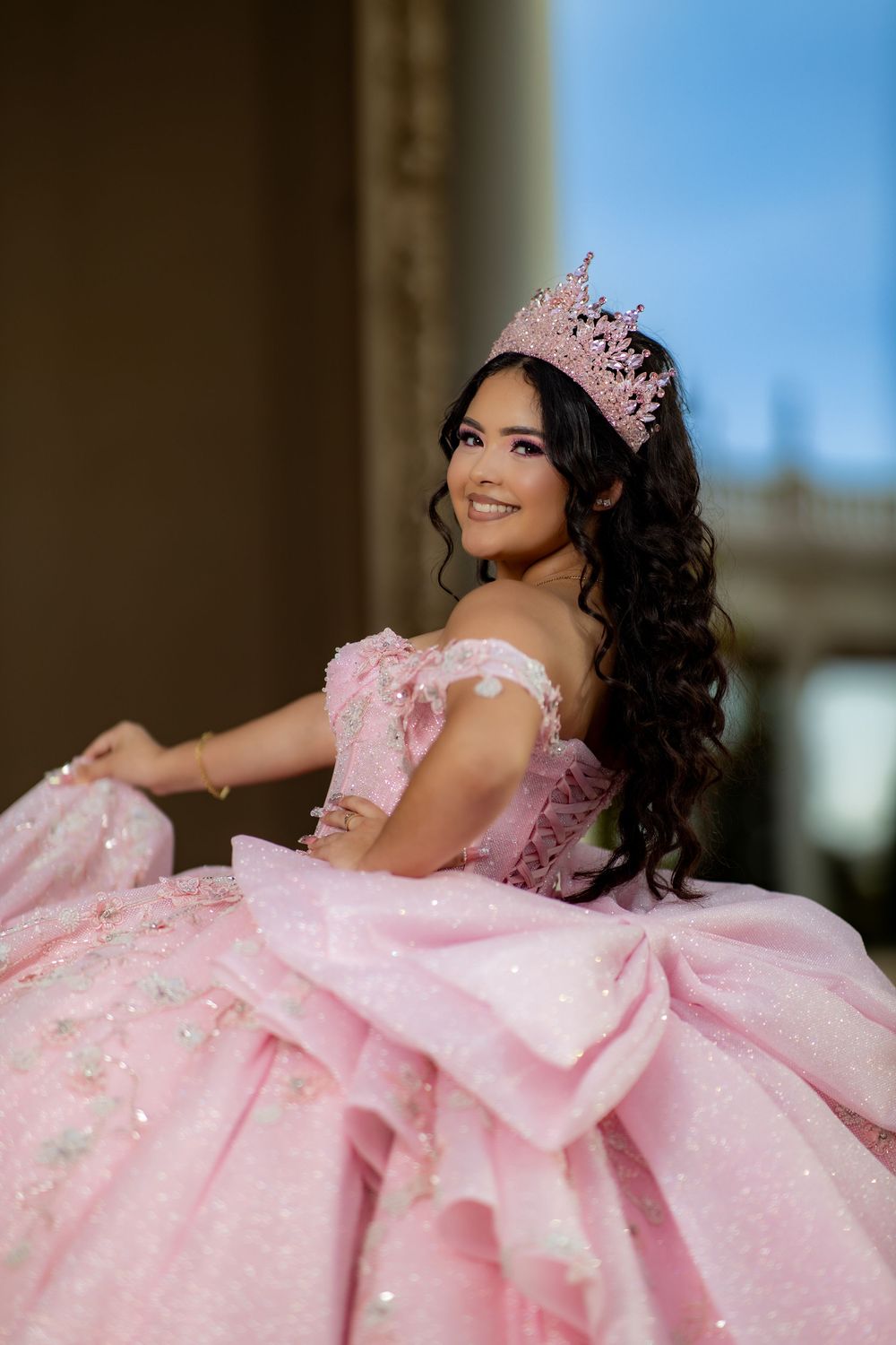 Smiling young woman in a sparkly pink quinceañera ball gown with a large bow and a rhinestone tiara, posing outdoors with long curly hair.