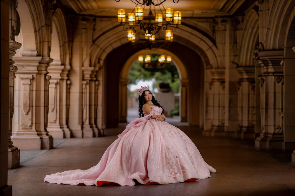 Young woman in a pink ball gown quinceañera dress poses elegantly in an ornate archway with chandelier.