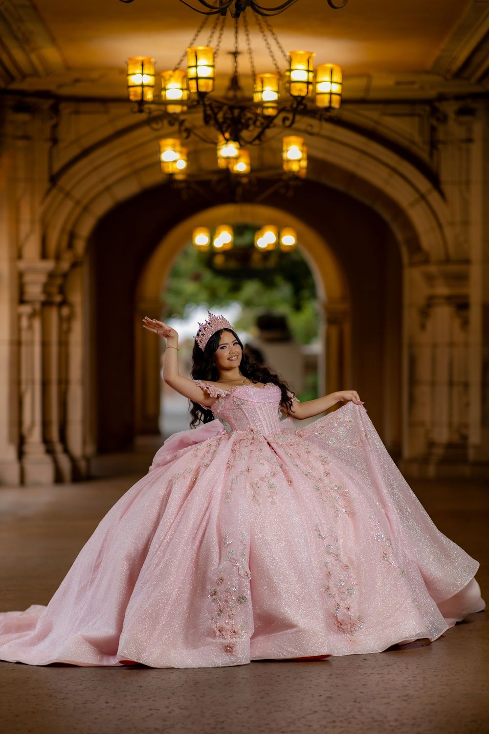 Woman in a sparkling pink quinceañera ball gown with floral appliqués and a tiara posing under a chandelier in an arched corridor.