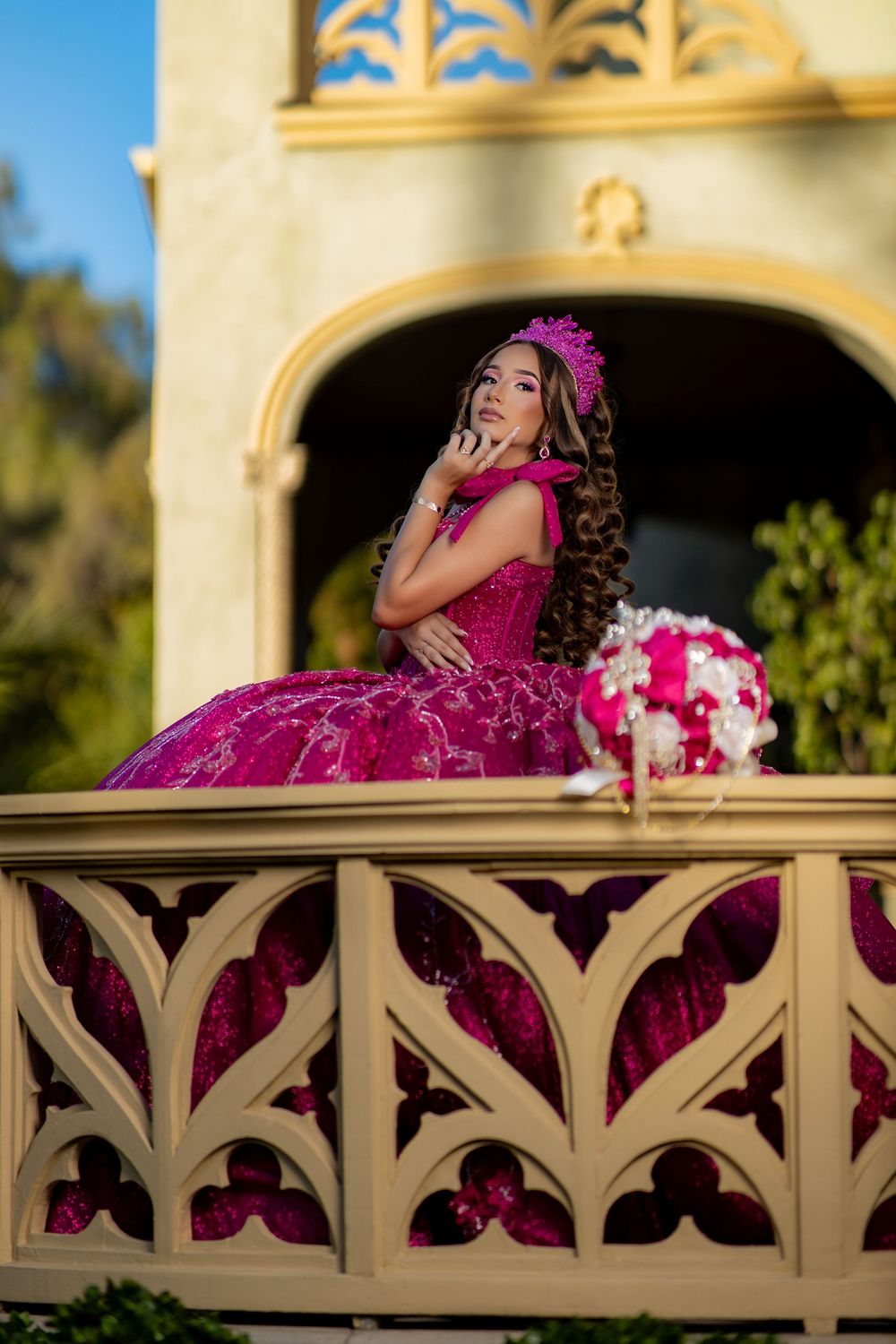 Young woman in a magenta quinceañera gown and crown poses gracefully on an ornate balcony with floral bouquet.