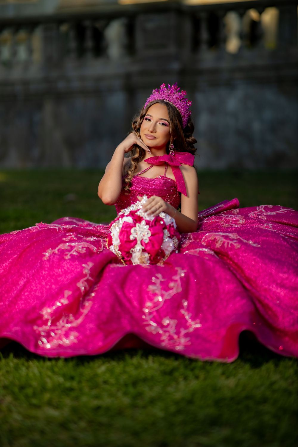 Young woman in a hot pink quinceañera ball gown and crown sits gracefully on green grass outdoors.