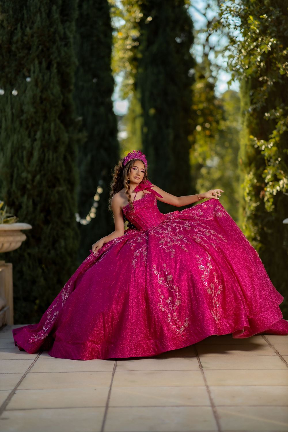 Young woman in a stunning fuchsia quinceañera ball gown with gold embroidery and floral crown poses outdoors.