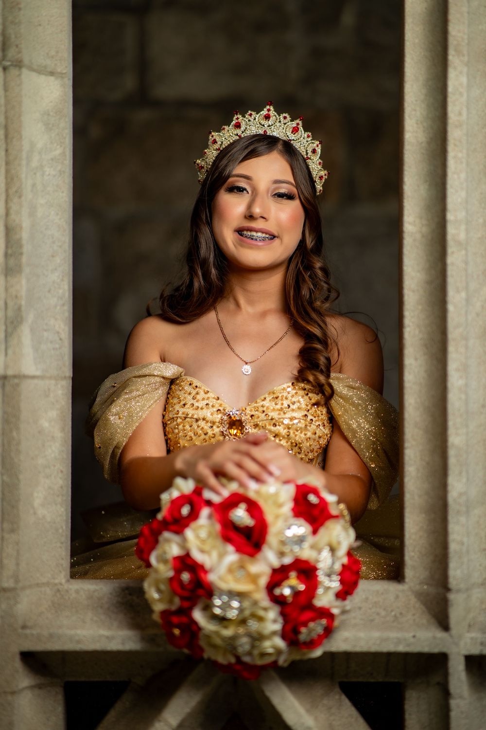 Young woman in gold sequin dress and red jeweled crown holds red and white rose bouquet at stone window.