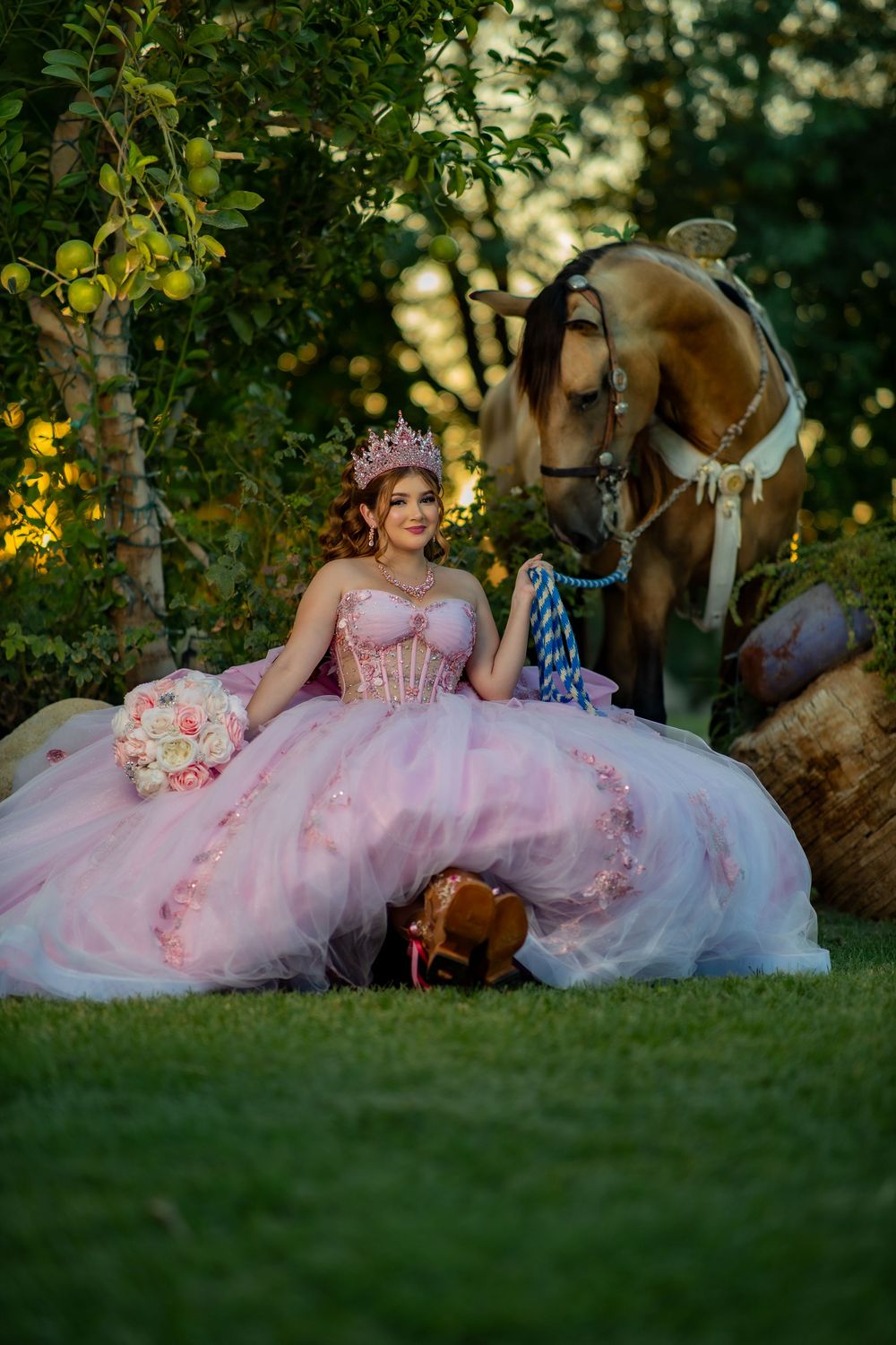Young woman in pink ball gown and crown sits beside a brown horse in a lush garden, holding a floral bouquet.