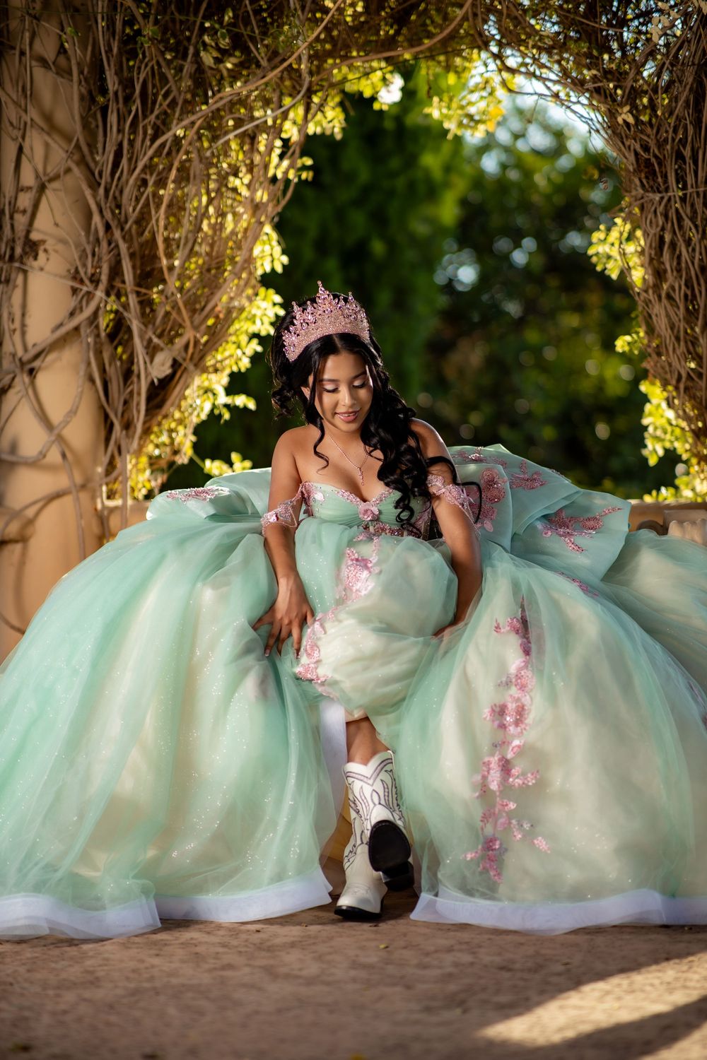 Young woman in mint green quinceañera ball gown with pink floral details and crown poses under vine arch outdoors.