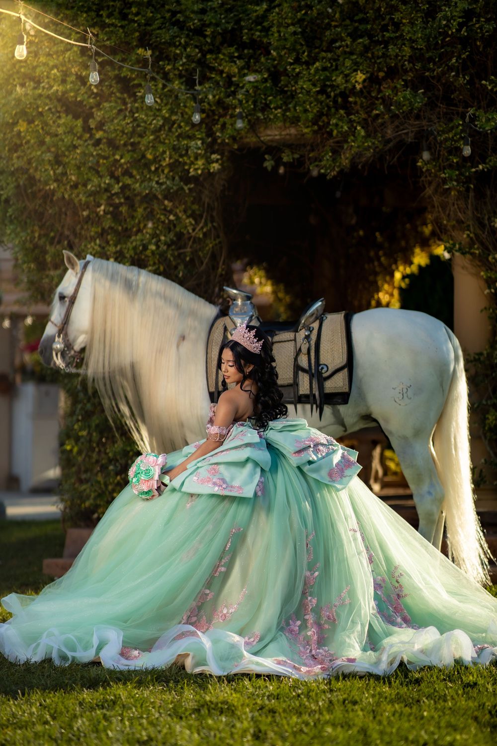 Young woman in mint green quinceañera gown with floral details poses beside a white horse outdoors.