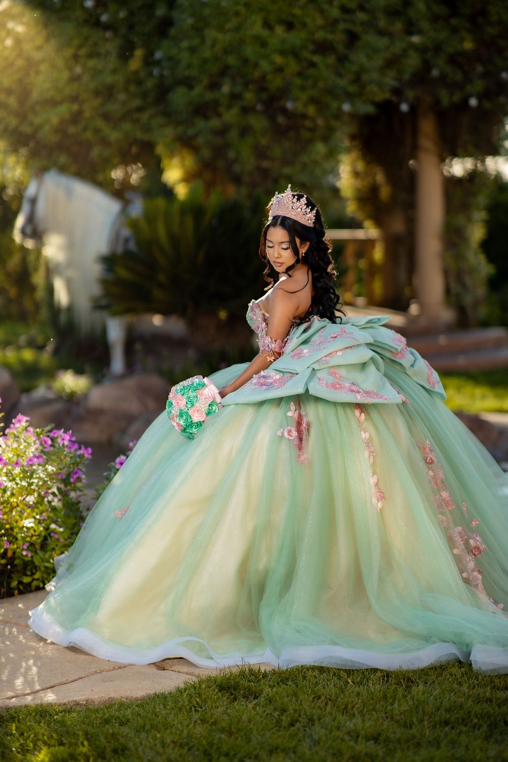 Young woman in a mint green quinceañera ball gown with floral details and tiara holds a bouquet in a garden.