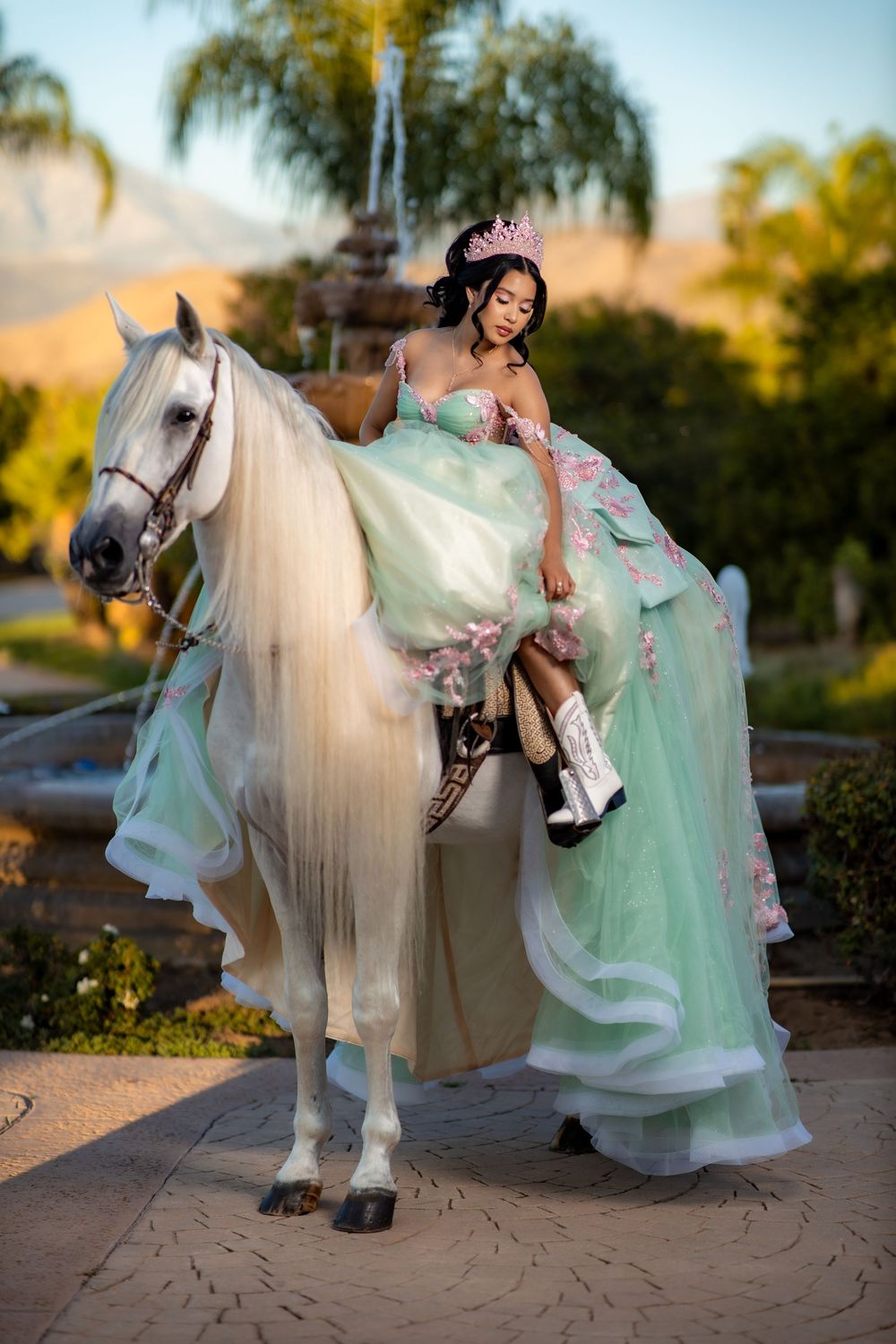 Young woman in mint green quinceañera gown and tiara sits elegantly on white horse outdoors at golden hour.