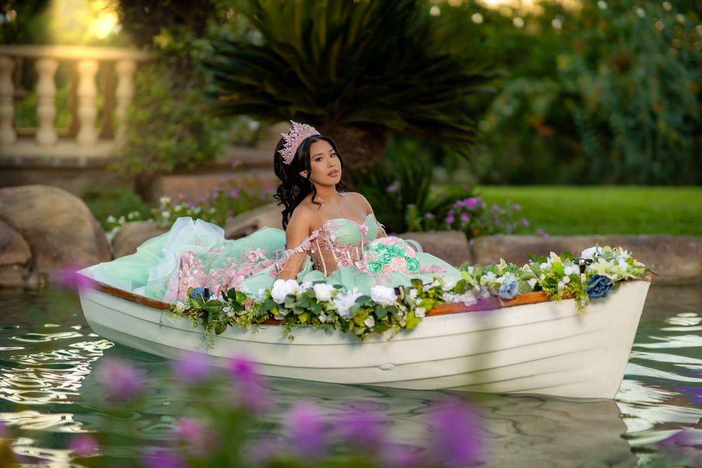 Young woman in mint green ball gown sits in flower-adorned white boat on serene garden pond.