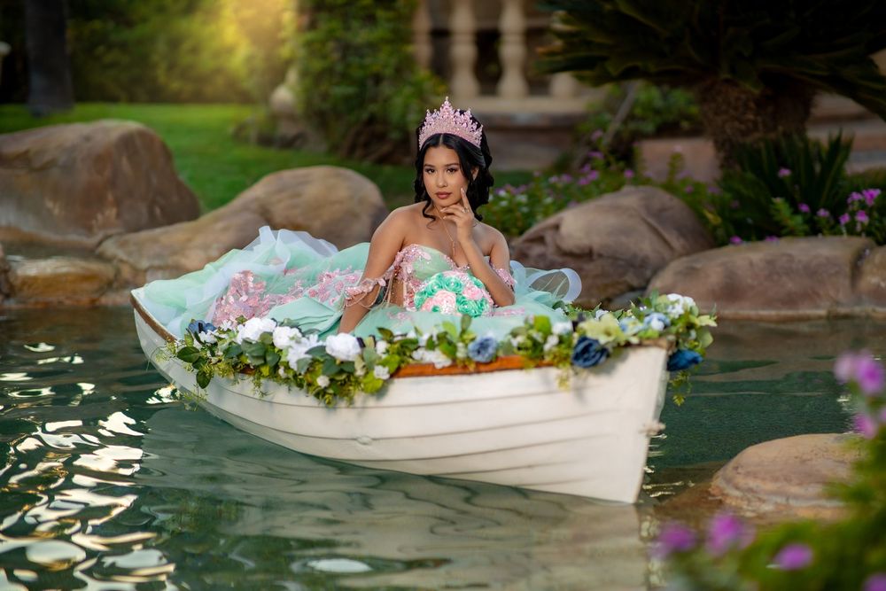 Young woman in a floral quinceañera gown and tiara sits in a white boat adorned with flowers on a serene pond.