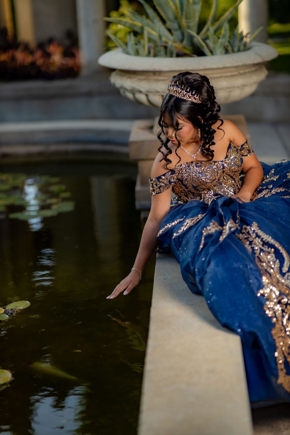 Young woman in a sparkling royal blue gown with gold embroidery and a tiara sits by a lily pond, touching the water with her hand.