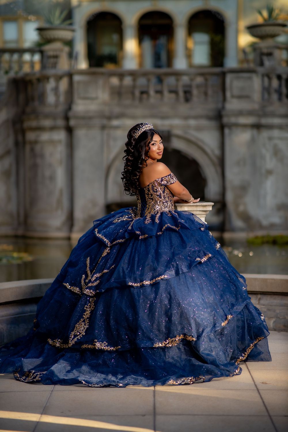 Young woman in a navy blue quinceañera ball gown with gold embroidery and a tiara, posing on a stone terrace beside a historic fountain.