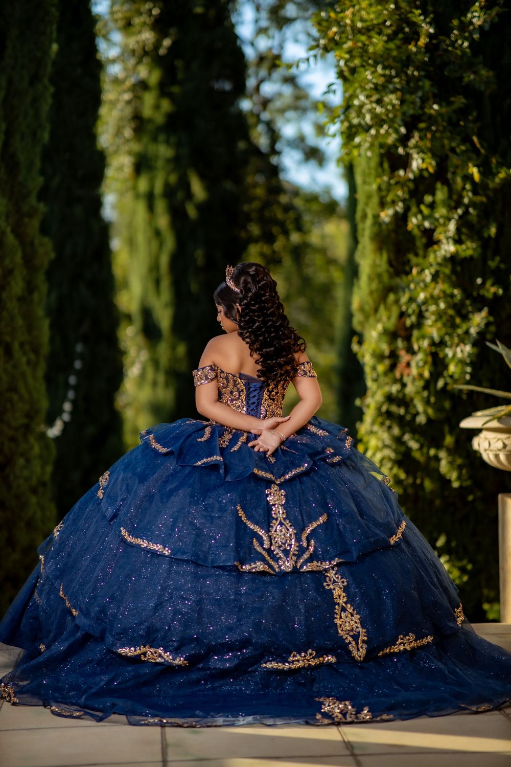 Woman in a navy blue off-shoulder quinceañera ball gown with gold embroidery and tiered tulle skirt, posed from behind in a garden.