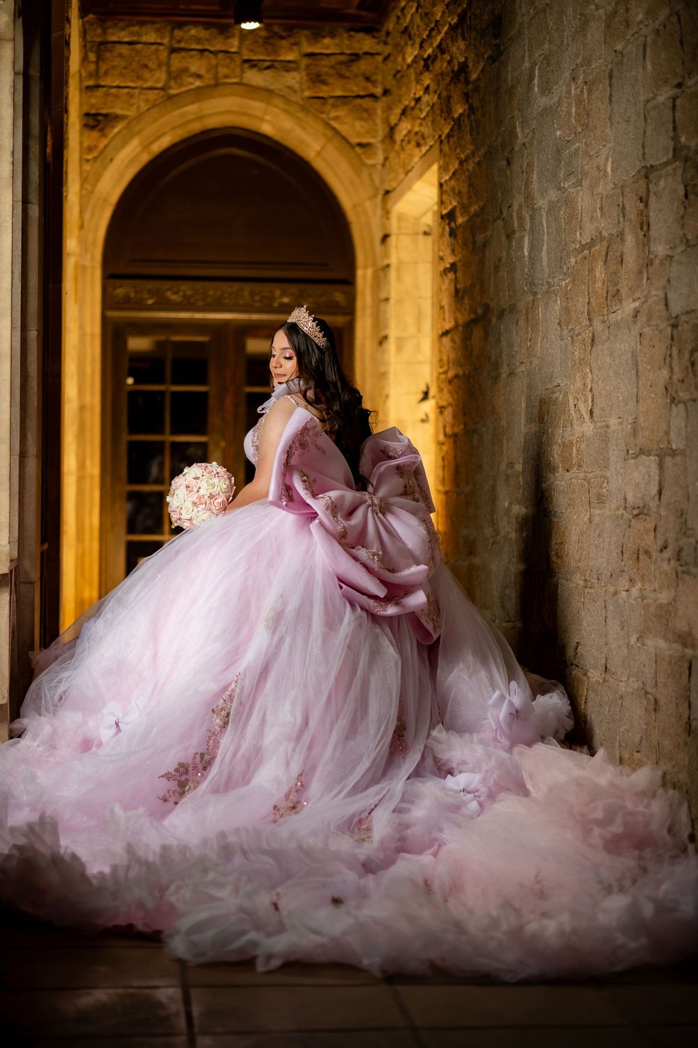 Young woman in pink ruffled quinceañera ball gown with large bow poses by stone archway at night.