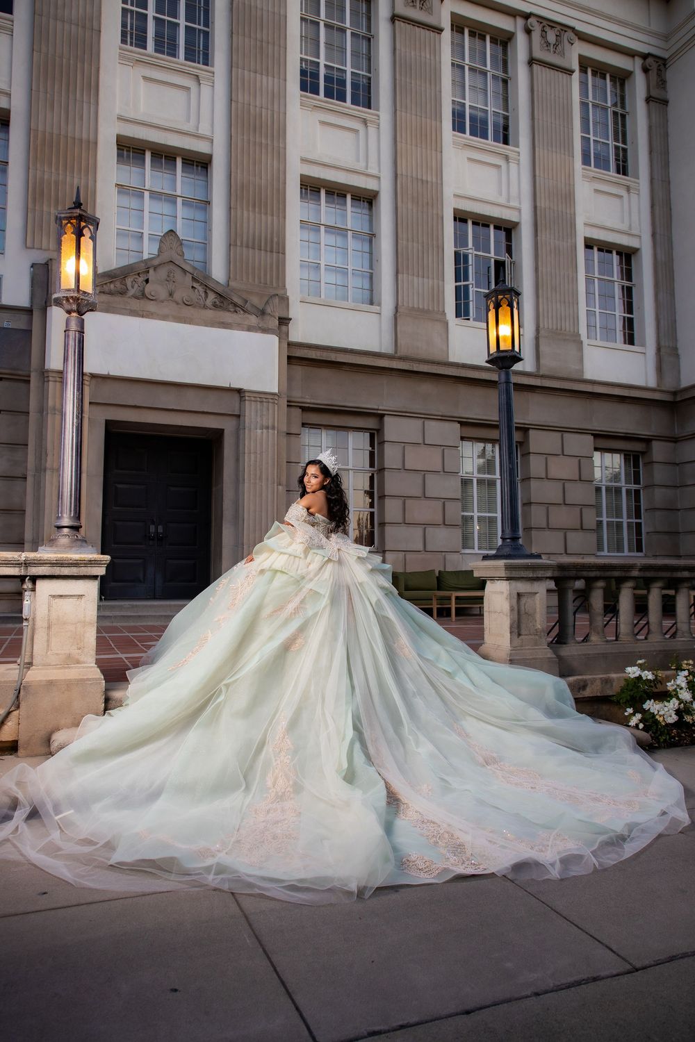 Woman in elegant mint green ball gown with floral details poses on steps of grand stone building at dusk.
