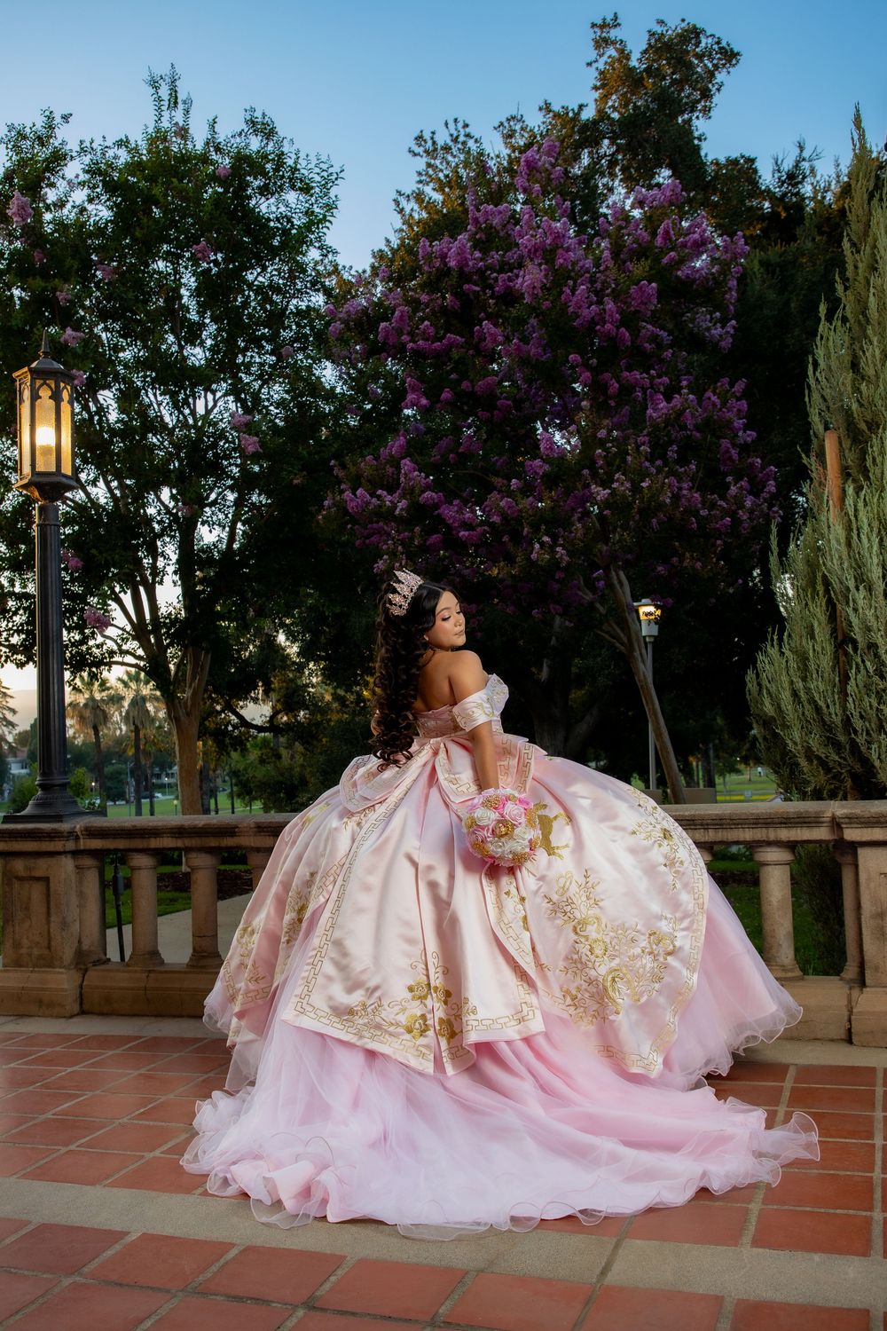 Young woman in pink ball gown with gold embroidery holds bouquet near blooming purple tree at elegant outdoor venue.