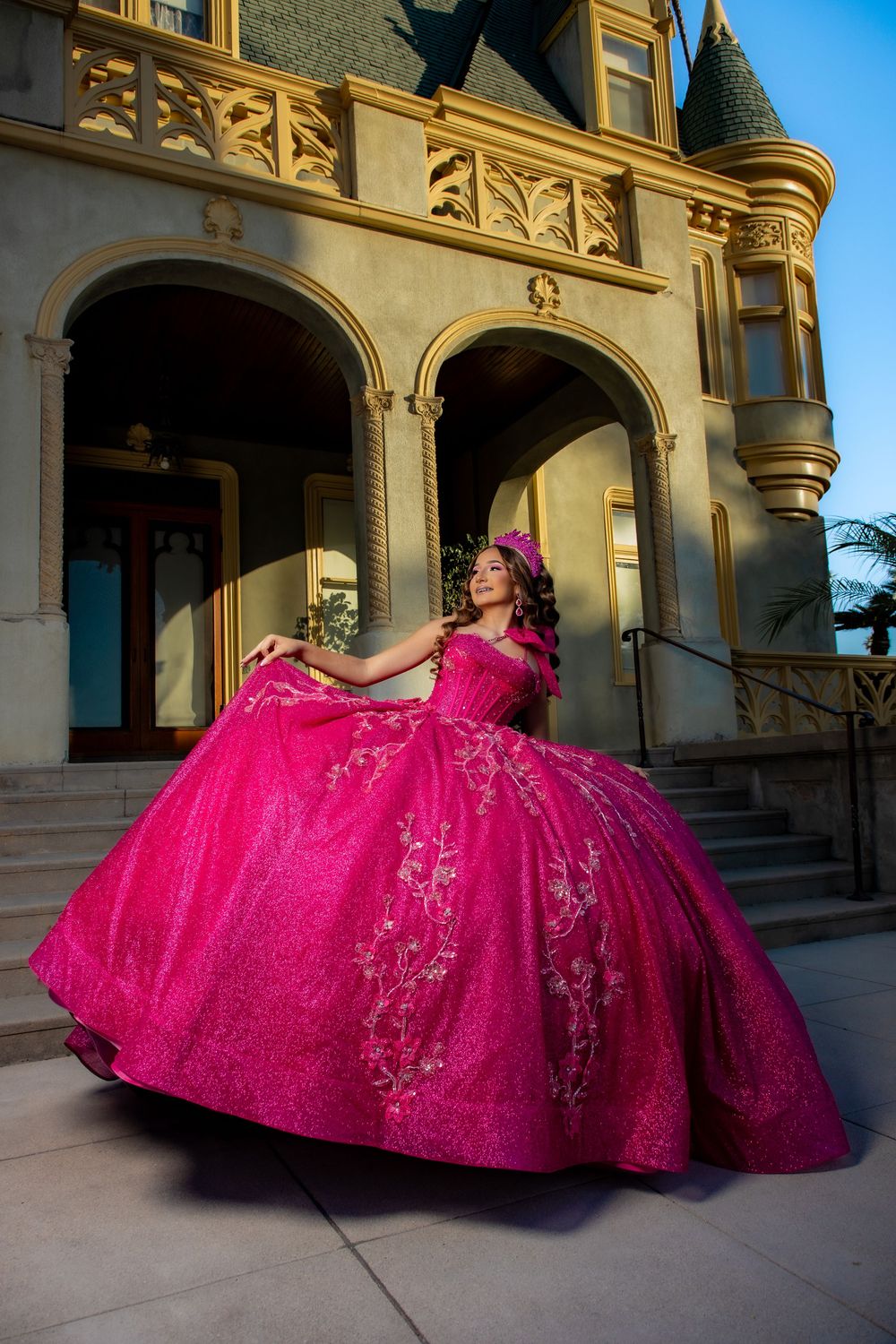 Young woman in a vibrant pink quinceañera ball gown with floral embroidery poses before a grand historic mansion.