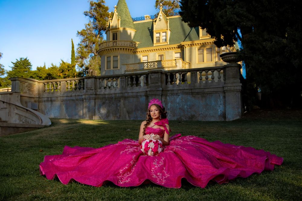 Girl in a voluminous hot pink quinceañera gown sitting on grass before a grand castle-like mansion.