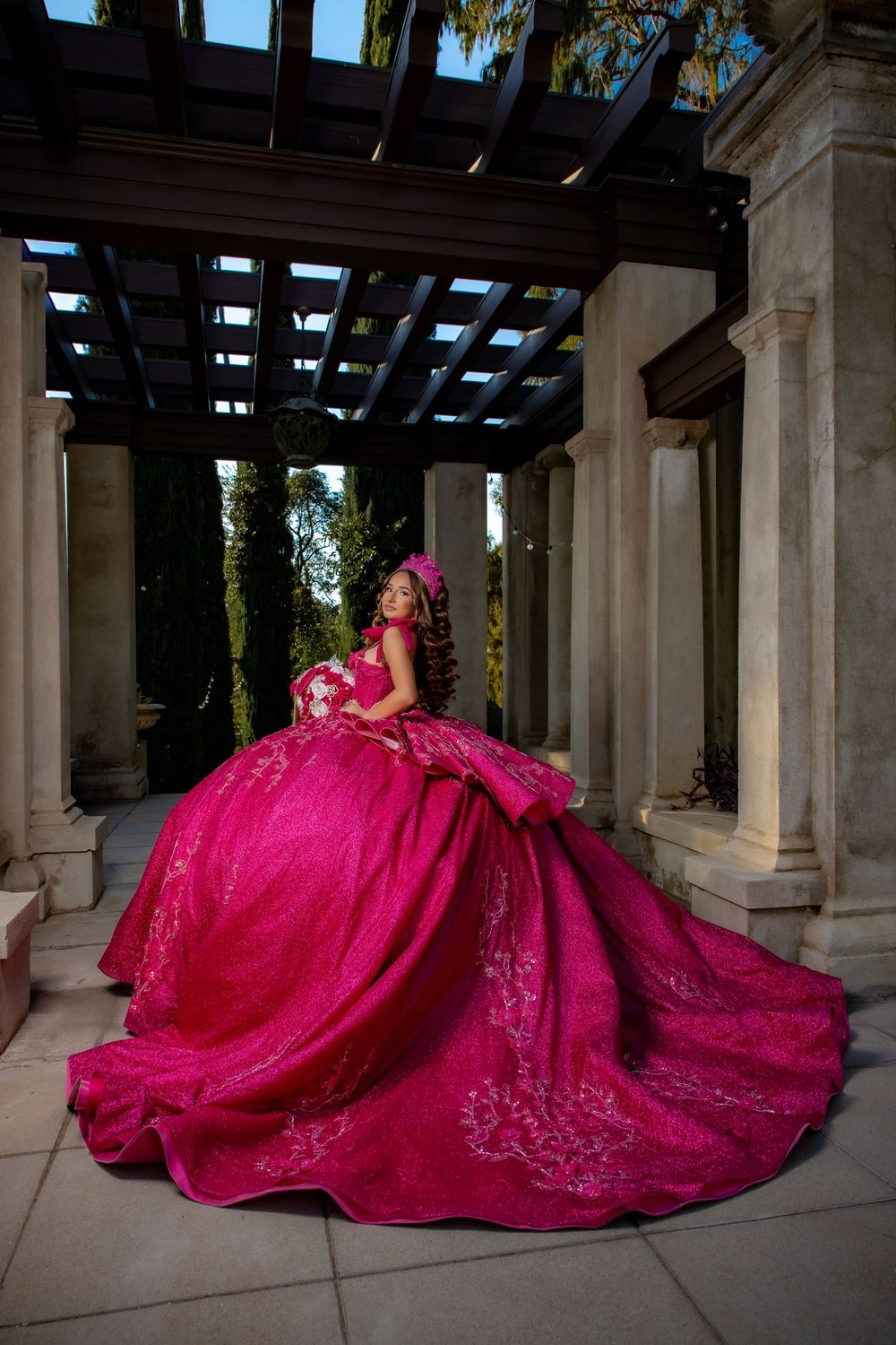 Young woman in a stunning fuchsia quinceañera ball gown with floral embroidery poses under an elegant pergola.