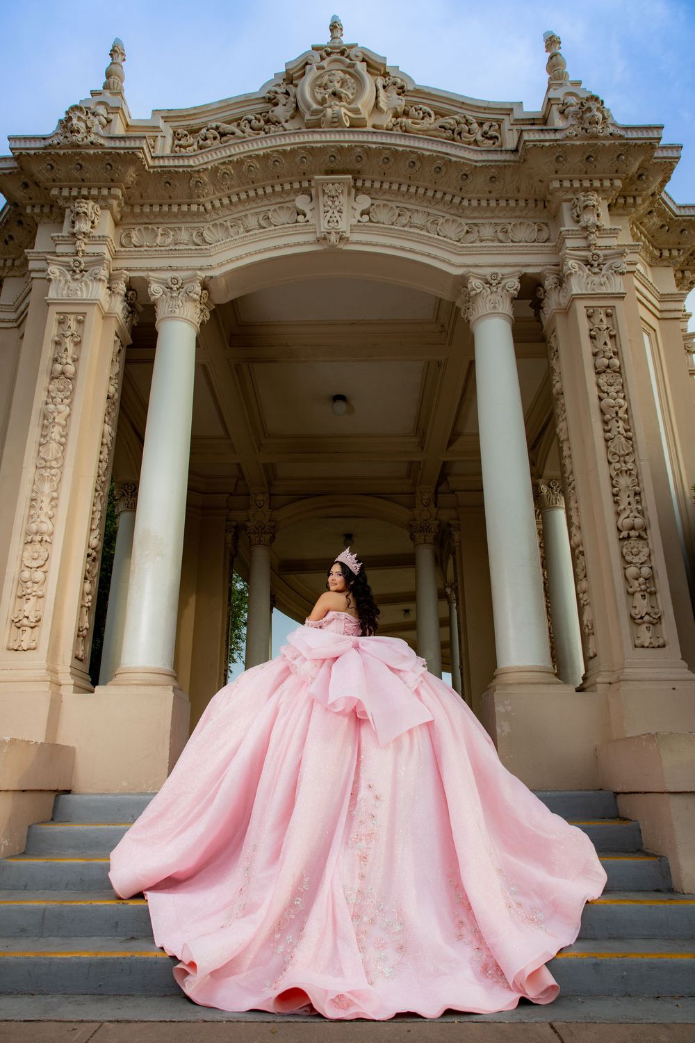 Young woman in a voluminous pink ball gown and crown poses on steps of an ornate neoclassical building.