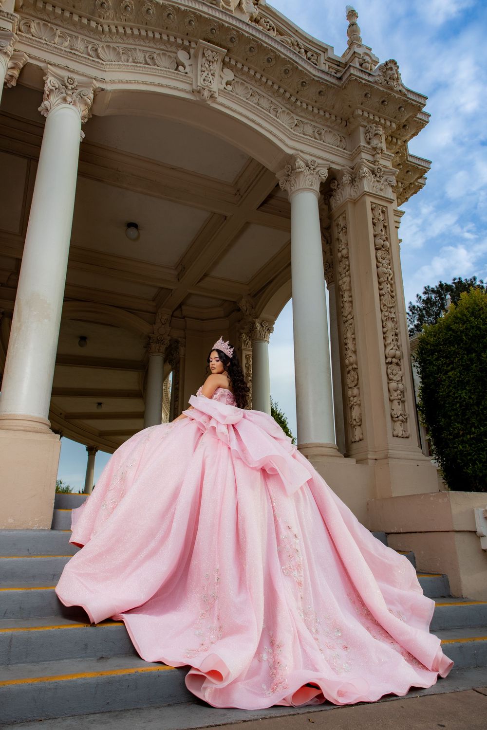 Woman in a pink quinceañera ball gown with a large bow and tiara posing on stone steps beneath an ornate columned pavilion.