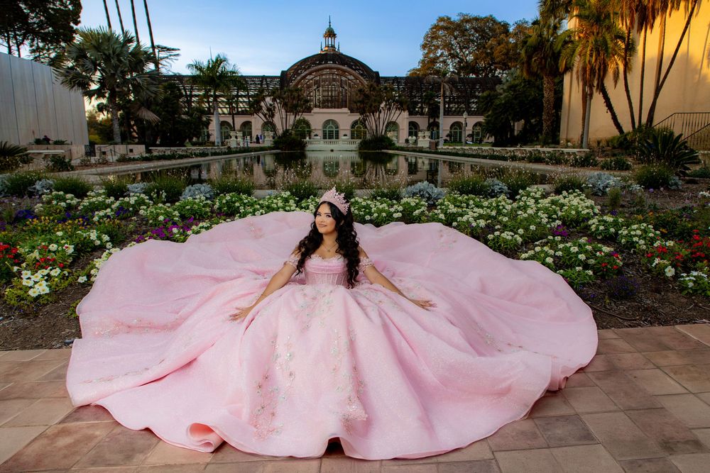 Young woman in a voluminous pink quinceañera ball gown and tiara posing on a tiled terrace beside a garden and reflecting pool.