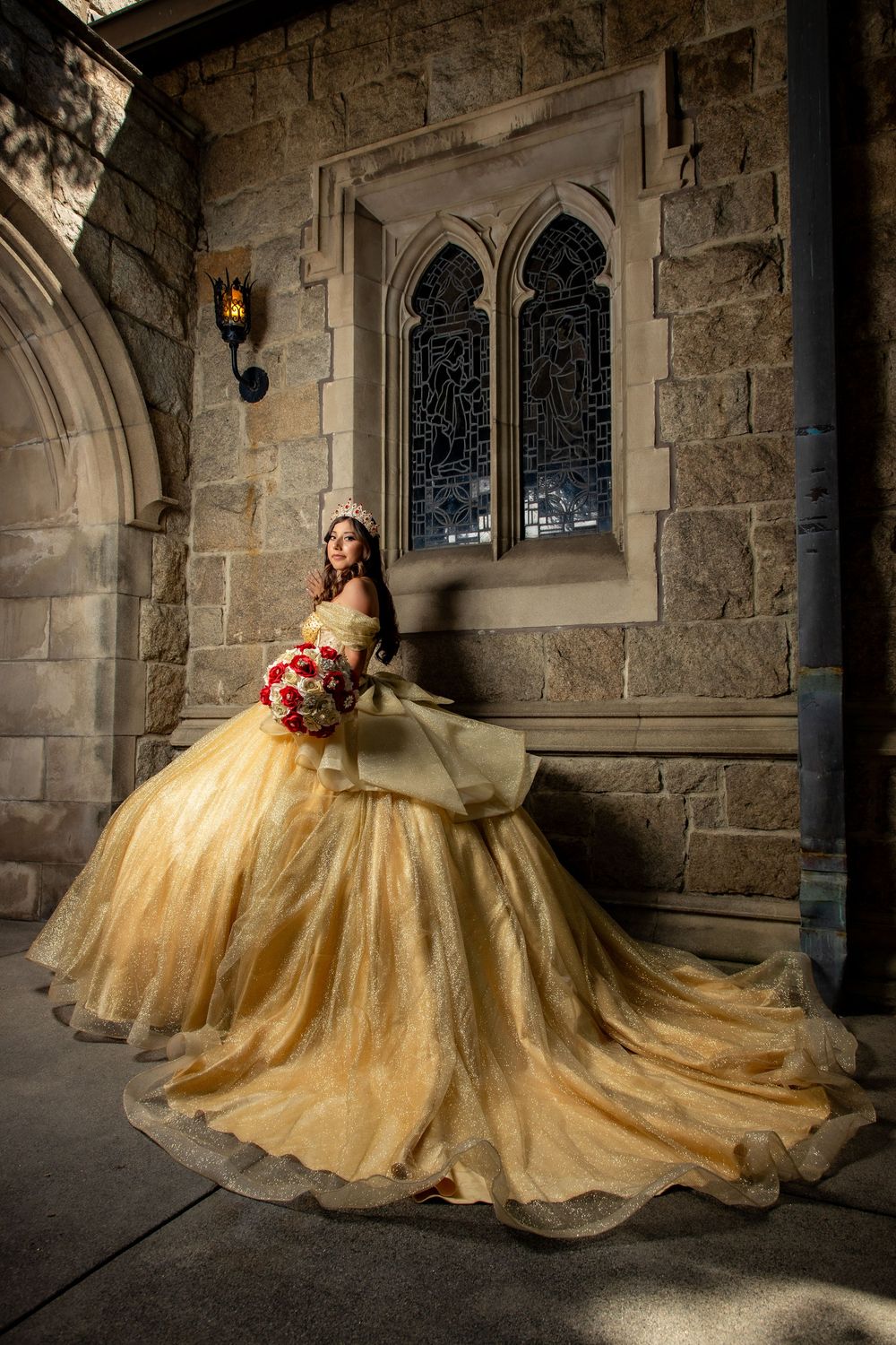 Young woman in golden ball gown holding bouquet poses elegantly before gothic stone castle architecture.