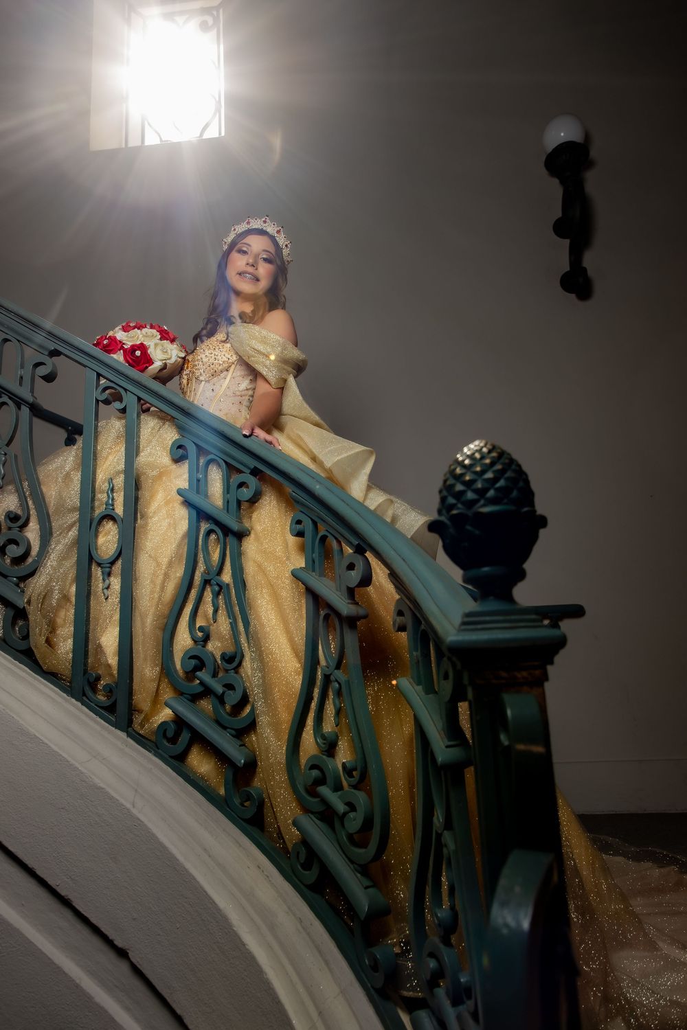Young woman in gold quinceañera gown with tiara holds bouquet on ornate teal staircase under dramatic lighting.