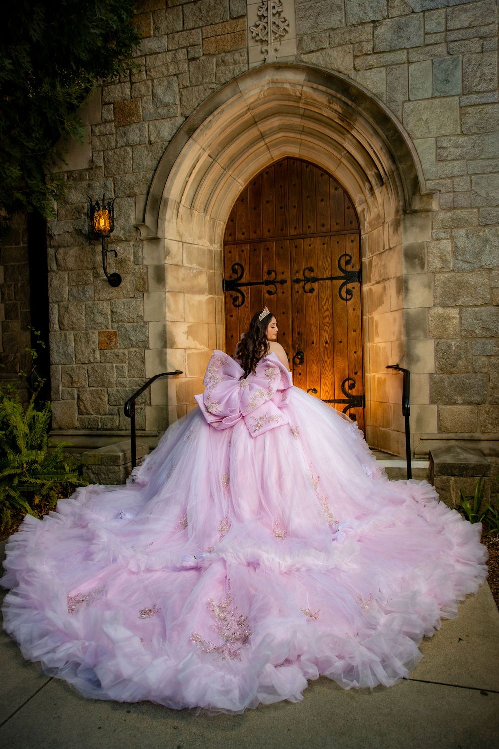 Girl in pink floral quinceañera ball gown with dramatic train stands before castle stone arch doorway.