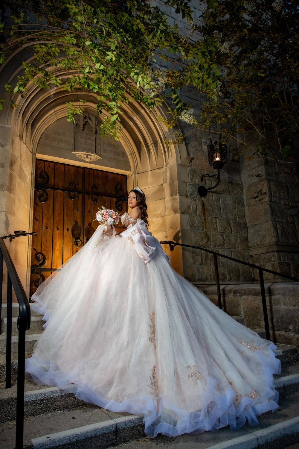Elegant bride in white gold-lace ball gown holds bouquet on illuminated stone church steps.