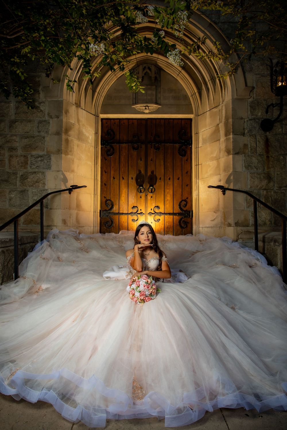 Bride in a voluminous blush ball gown sits on church steps holding a floral bouquet before ornate wooden doors.