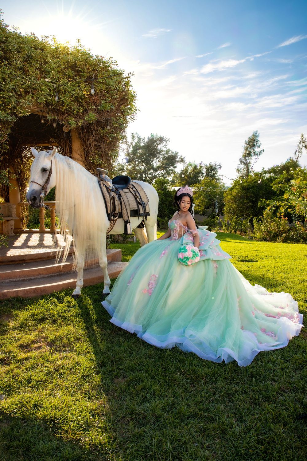 Young woman in mint green quinceañera ball gown poses beside a white horse in a sunlit garden setting.
