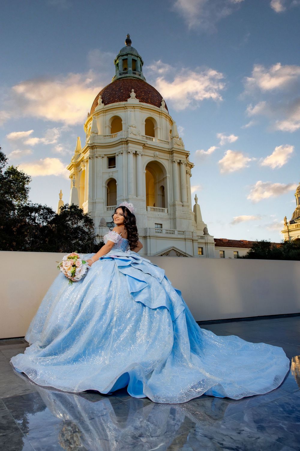 Quinceañera girl in blue sequined ball gown posing before Pasadena City Hall at sunset.