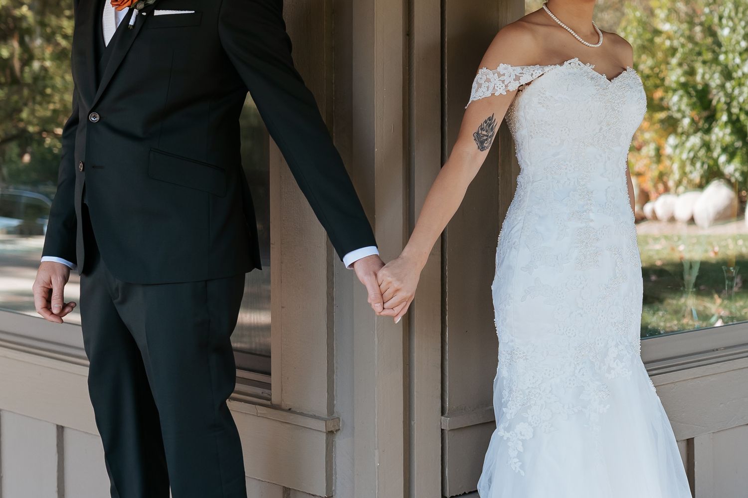 Couple in wedding attire holding hands while standing on opposite sides of a building column outdoors.