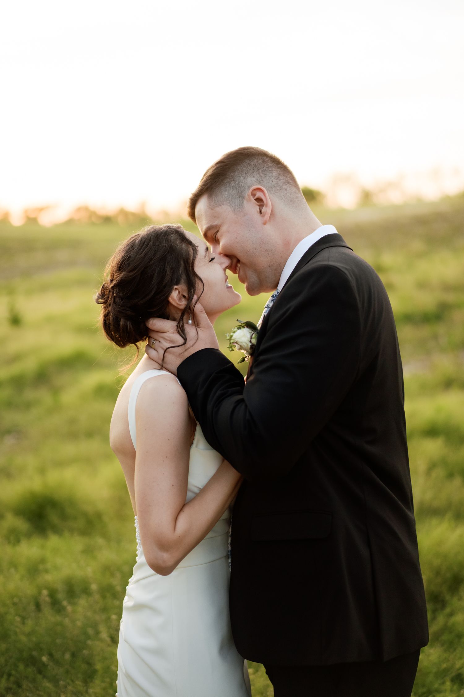Newlywed couple walking together in a sunlit meadow with bride in flowing white dress and groom in dark suit.