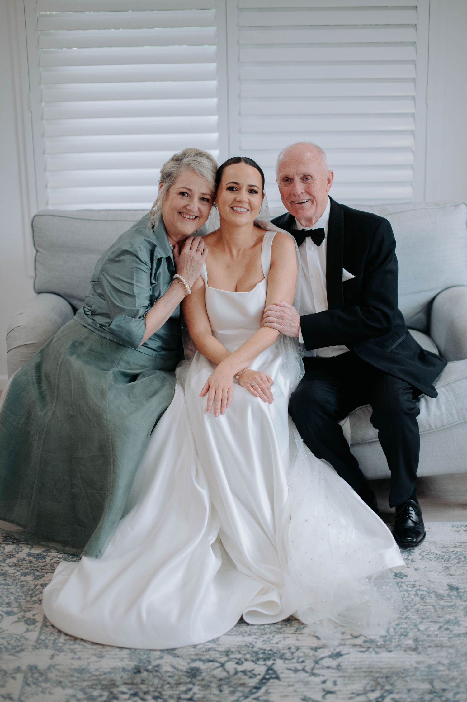A bride in a white satin gown sits on a gray couch with two guests at a formal wedding celebration.