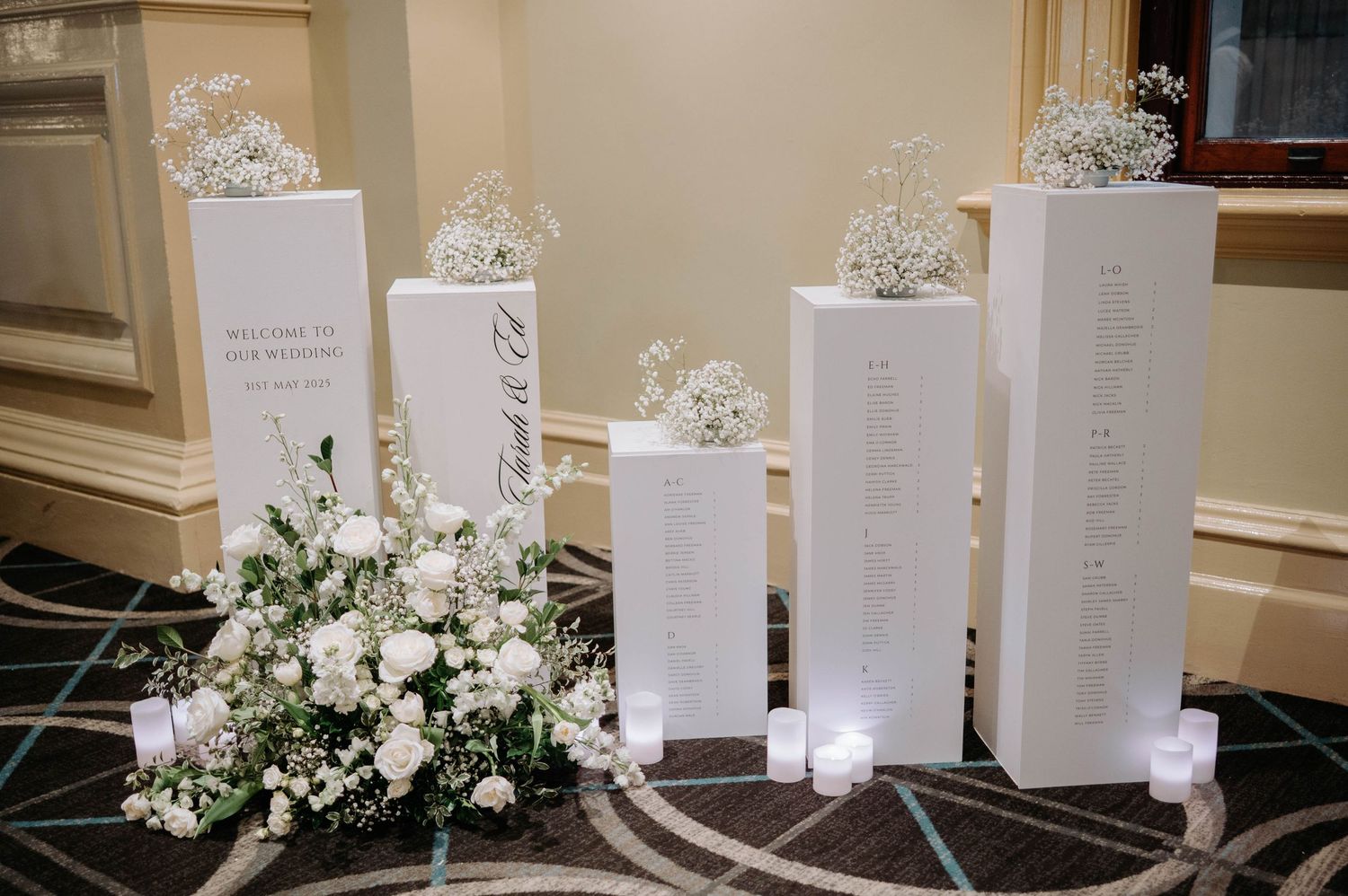 White wedding seating chart displays on illuminated columns with floral arrangements at the base against beige wall decor.