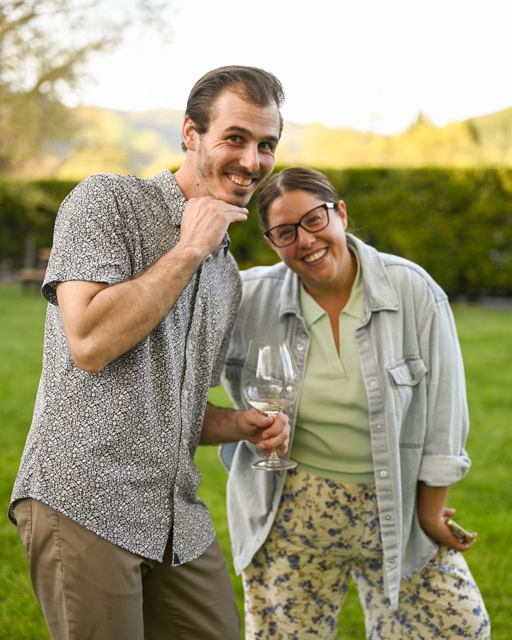 Two friends share a laugh and toast with wine glasses while enjoying a sunny outdoor gathering.