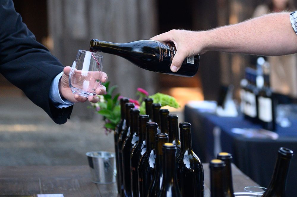 A wine pourer serves guests at a tasting event in Napa Valley, surrounded by dark wine bottles at Reid Family Vineyard.