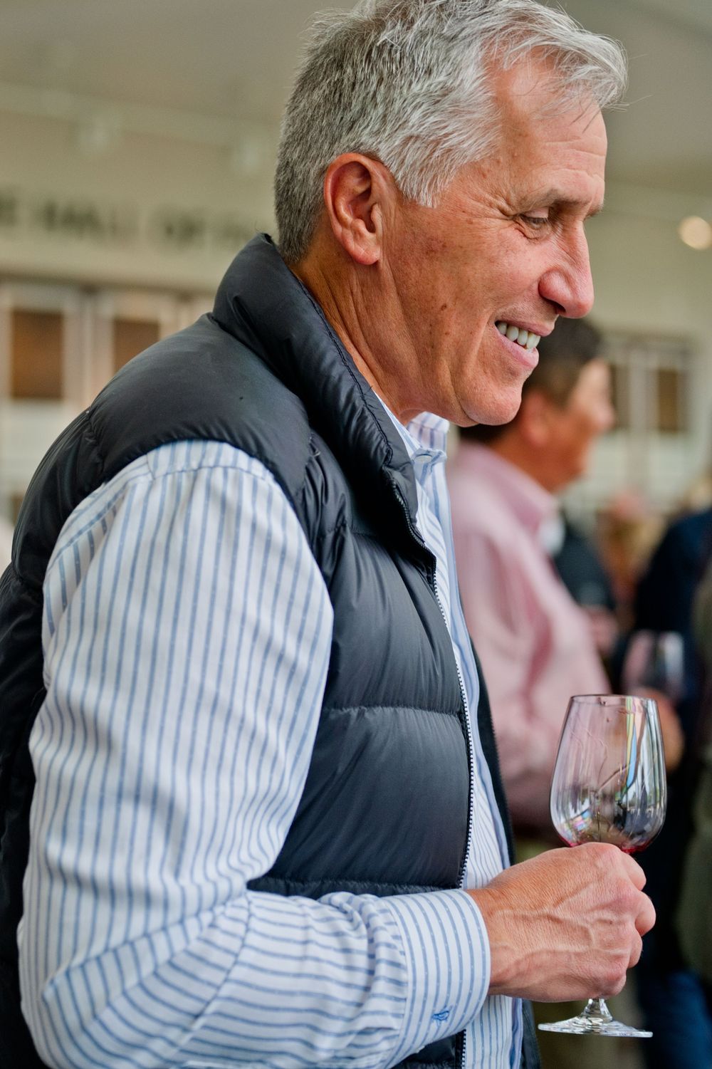 A person in a black puffy vest and striped shirt smiles while holding a wine glass during a social gathering.