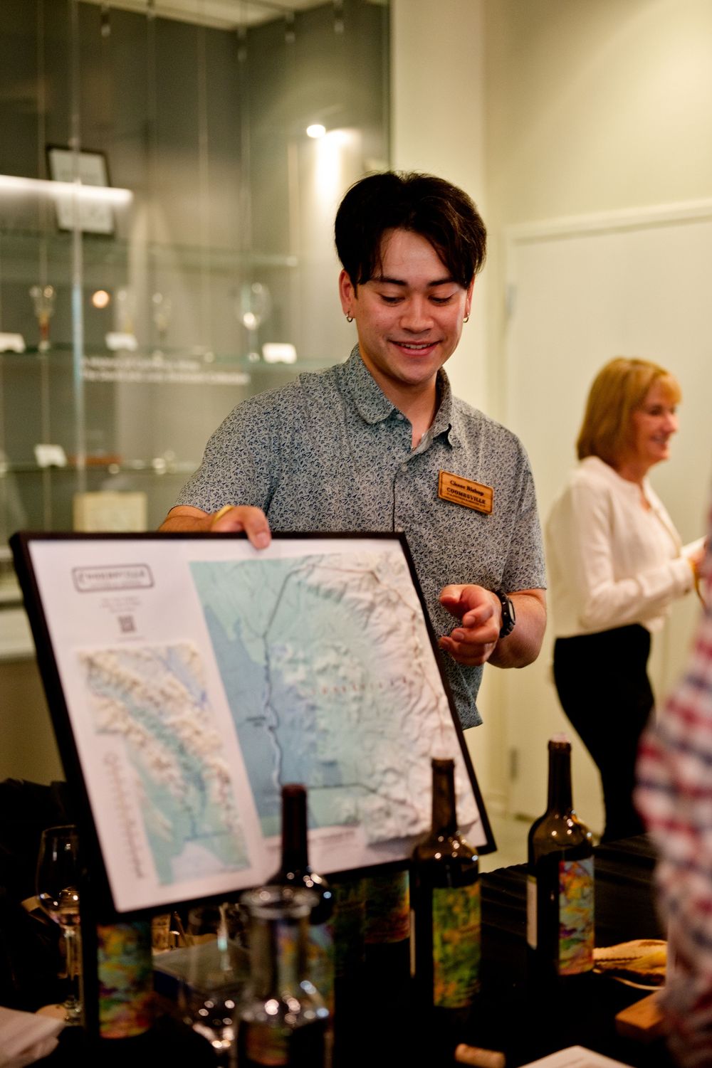 Wine steward smiles while presenting a map display at a wine tasting event with bottles arranged on the table. CIA Copia - Napa, CA