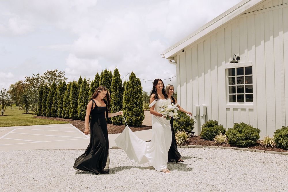 Bride in white gown walks with two bridesmaids in black dresses outside white barn venue.