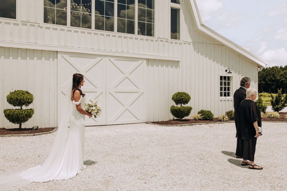 Bride in fitted white gown holds bouquet during first look outside white barn wedding venue.