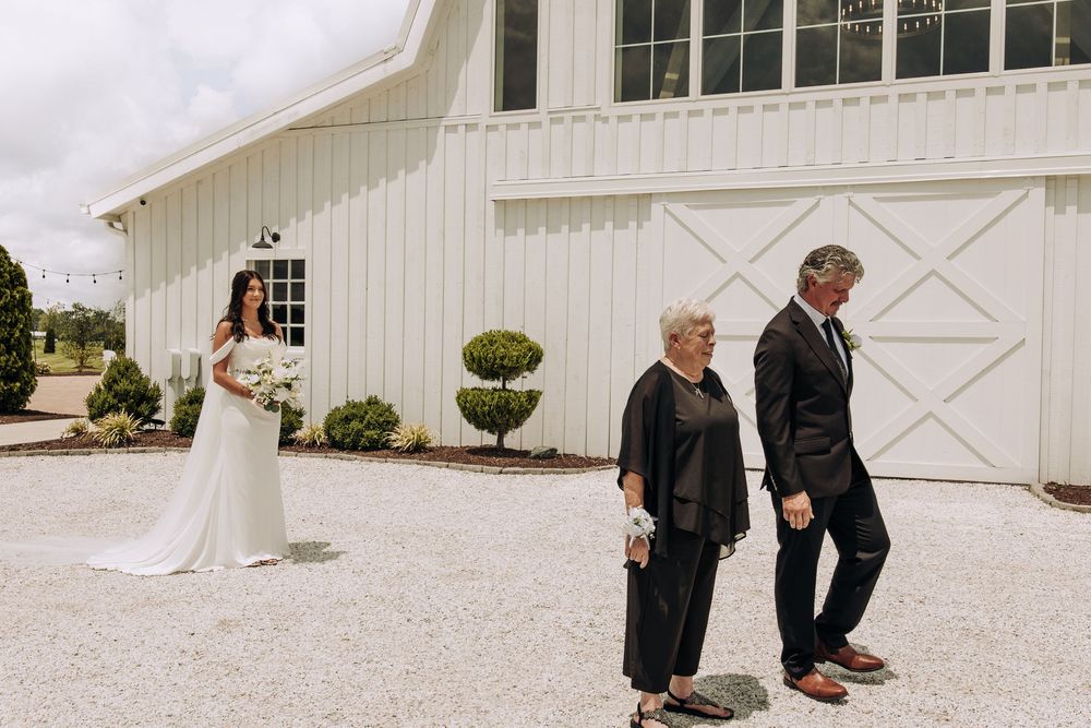 Bride stands before parents during emotional first look outside elegant white barn venue.