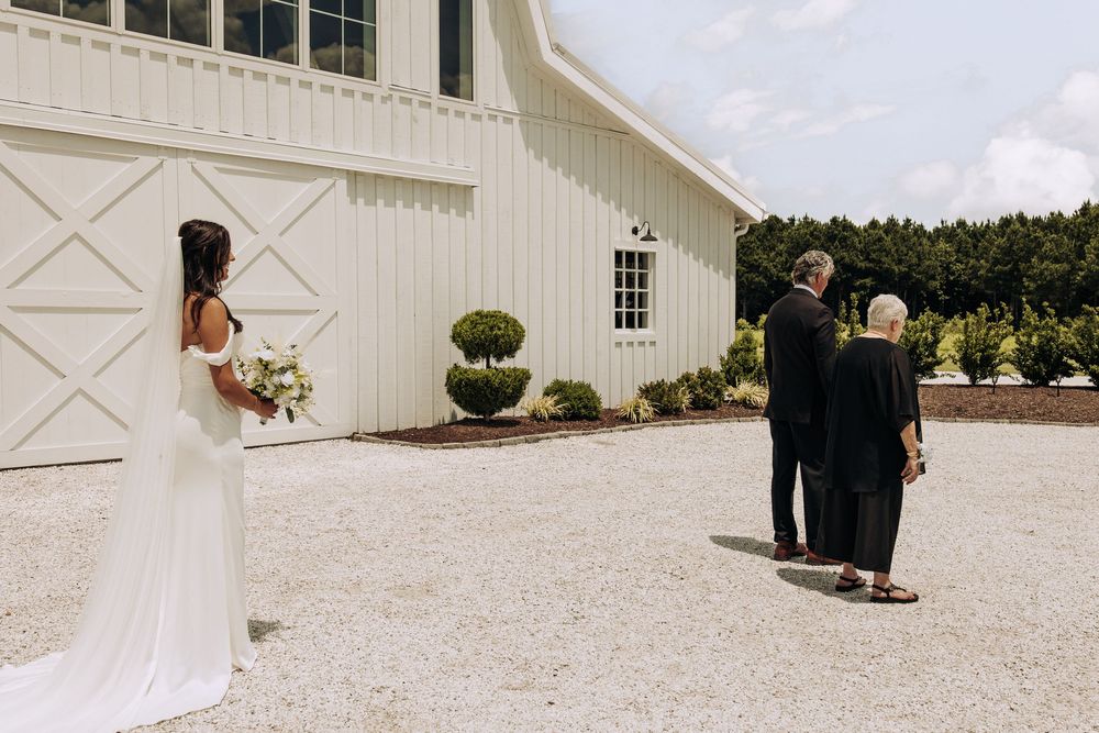 Bride in white gown faces parents during outdoor first look at white barn wedding venue.