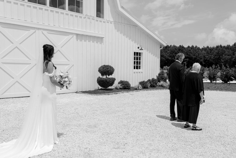 Black and white photo of bride sharing first look moment with parents outside white barn.