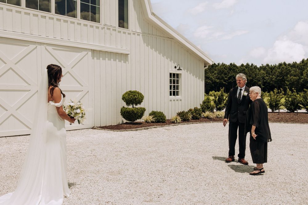 Bride in elegant white gown shares emotional first look with parents at barn wedding venue.